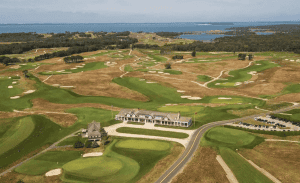 Aerial view of a sprawling golf course near a body of water. The landscape features green fairways, sand bunkers, and water hazards. A clubhouse with adjacent smaller building and parking lot is situated near the course. Rolling hills and trees are scattered throughout—ideal for property management in East Hampton.