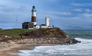 A scenic view of a lighthouse perched on a grassy cliff overlooking the ocean. The lighthouse is flanked by smaller buildings, and the rocky shoreline below is met by waves crashing against it. The sky is partly cloudy, adding to the picturesque coastal scene reminiscent of Property Management East Hampton's finest locations.