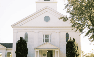 A white historic church with a gabled roof, arched windows, and a small circular window near the top. The entrance features a pediment supported by columns, flanked by tall shrubs. Tree branches partially frame the right side of the image. Ideal for those considering Rental Property Management in East Hampton.