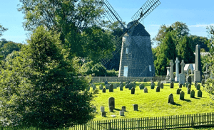 A historic windmill stands behind an old cemetery on a sunny day. The cemetery, enclosed by a wooden fence, has numerous headstones of varying sizes. Tall trees and lush green grass are seen throughout the landscape, creating a peaceful setting in East Hampton—a charming area known for its luxury home management services.