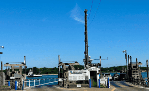 Image showing a ferry dock on a clear day. A ferry is loading vehicles onto its deck. Signs, an ATM, and barriers are visible at the dock entrance. There are trees and a body of water in the background, perfectly capturing the charm that makes Property Management East Hampton so desirable.