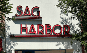 Image of the Sag Harbor Theater sign. The words "SAG HARBOR" are displayed in large, red, vintage letters on a white wall. The sign is bordered by trees, casting shadows on the building, evoking the charm that makes East Hampton's property management so desirable.