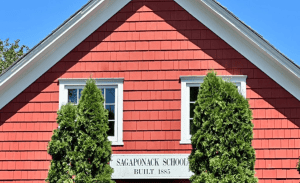 A historic red building with a steep gabled roof and white trim, labeled "Sagaponack School, Built 1885" on a sign beneath two windows. Two tall evergreen trees partially obscure the sign. Clear blue sky in the background, evoking the charm perfect for Rental Property Management East Hampton specialists.