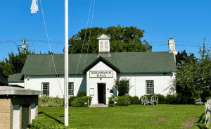 A white building with a green roof, identified as Ashawagh Hall by the sign above the entrance, is set against a bright blue sky. The building has a small cupola and is surrounded by well-maintained green lawns and trees. A flagpole stands in the foreground, showcasing East Hampton's charm.