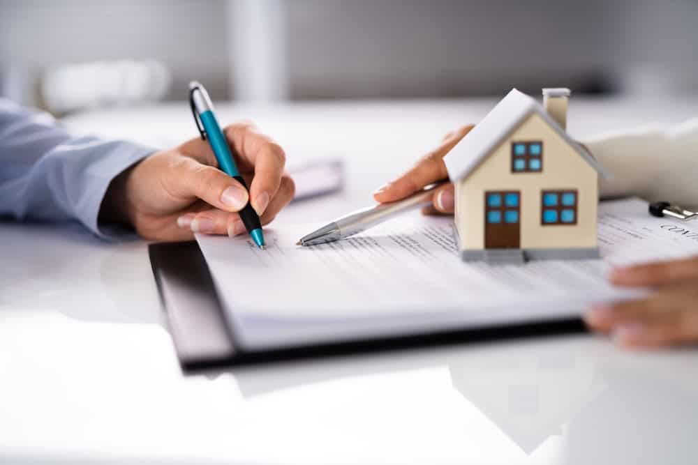 Two individuals are signing a document on a clipboard. One holds a pen while the other points to the paper. A small, model house sits on the table near the clipboard, indicating the document pertains to luxury home management in East Hampton.