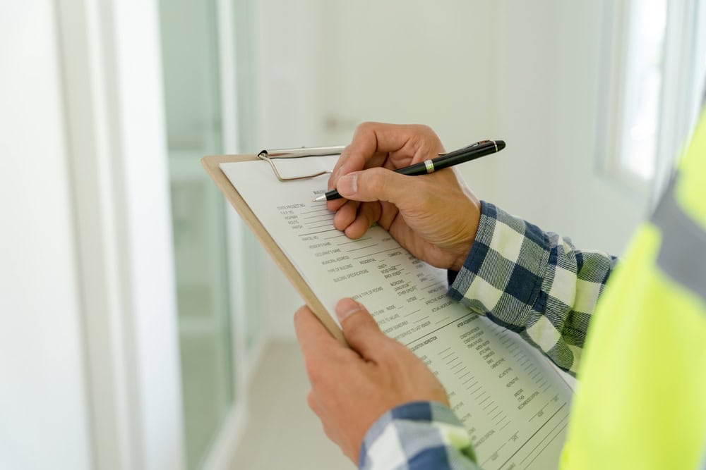 A person in a checkered shirt holds a clipboard with a document, using a pen to fill out or check information. The individual appears to be in a bright indoor setting, possibly overseeing Luxury Home Management East Hampton in a workplace or building inspection environment.