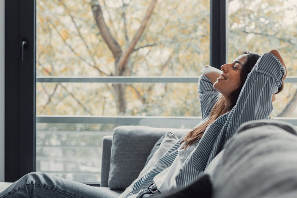 A woman is sitting relaxed on a gray couch with her hands behind her head and eyes closed, smiling. She is wearing a striped shirt and jeans. Behind her, a large window reveals a view of trees with autumn leaves. The room appears bright and tranquil, reflecting the calm brought by Luxury Home Management East Hampton.