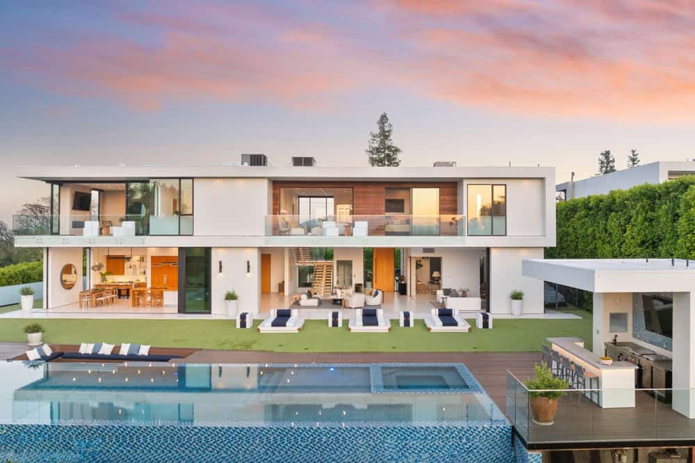 A modern, two-story luxury mansion with large glass windows and an open-concept design, managed by Rental Property Management East Hampton. The spacious backyard features a well-manicured lawn, multiple lounge areas, an outdoor dining area, and an infinity pool with a surrounding deck. The sky shows a beautiful sunset.