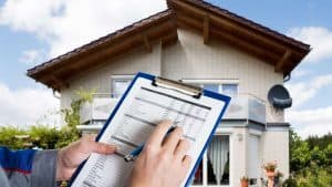 A person holding a clipboard with a checklist is standing in front of a two-story house. The house has white brick walls, a sloped roof, and a satellite dish. The sky is partly cloudy.