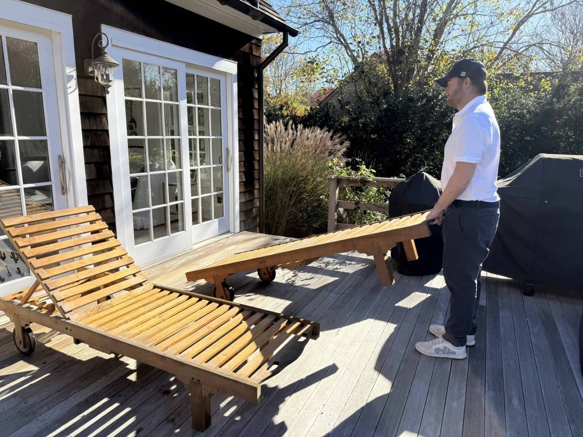 A person in a white shirt and cap adjusts a wooden lounge chair on a sunlit deck with a glass door and railing. Two covered grills are visible nearby. Tall grasses and trees are in the background.