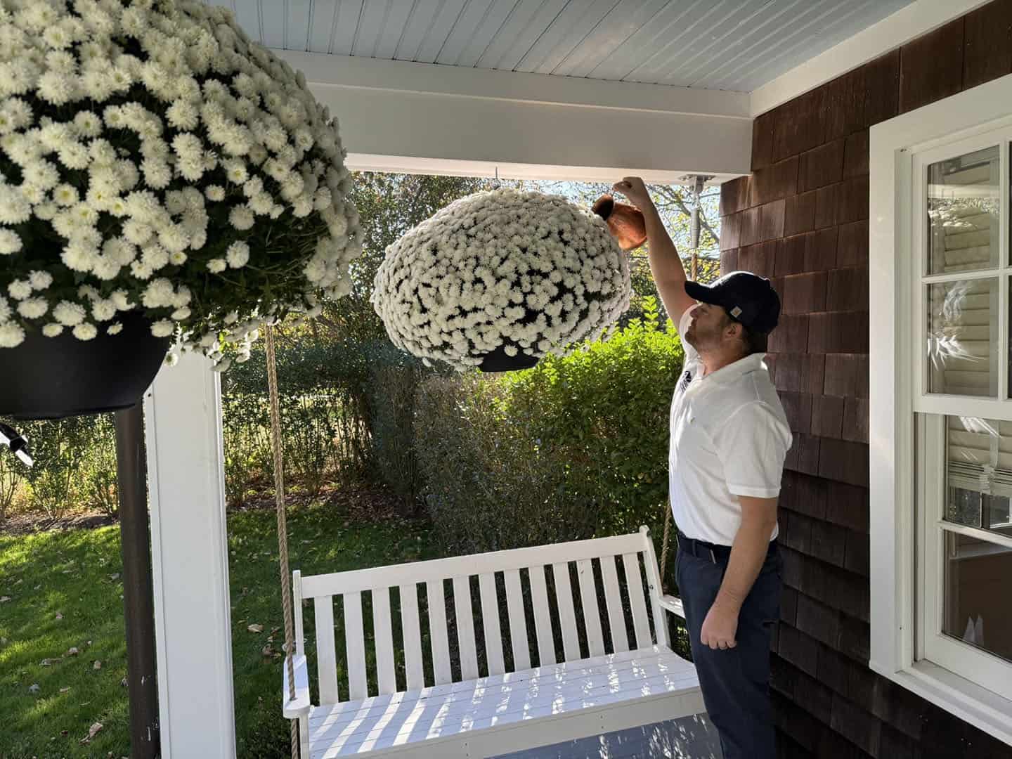 A man in a white shirt and black cap waters large hanging baskets of white flowers on a porch. He stands by a white swing bench, surrounded by green foliage in the background.