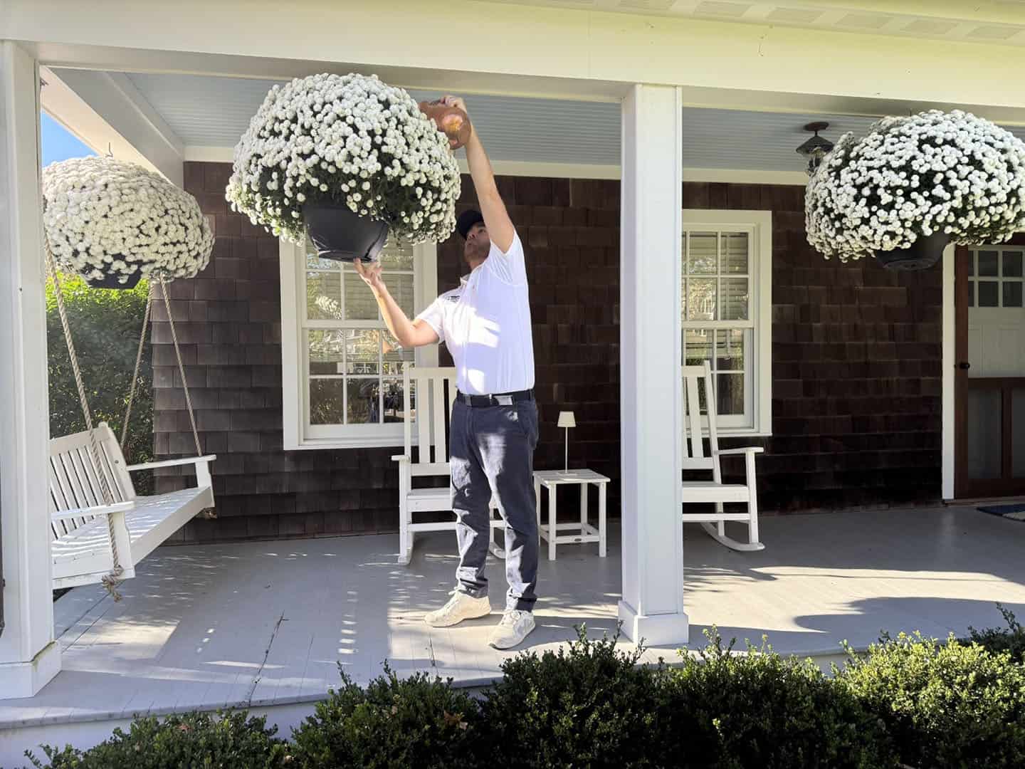 A person in a white shirt and cap stands on a porch adjusting a hanging basket of white flowers. The porch features a swing and rocking chairs, with sunlight casting shadows on the wooden floor.