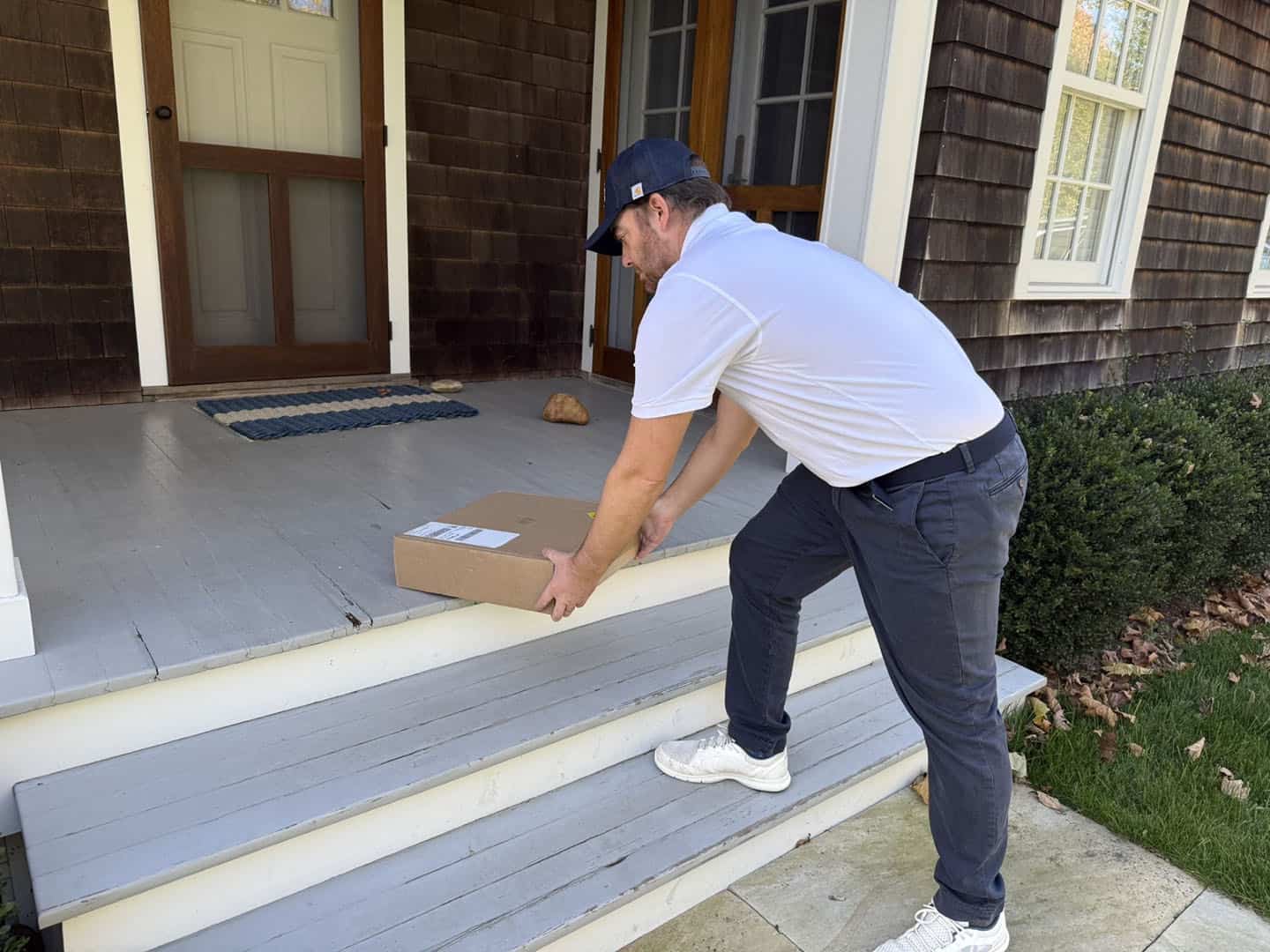 A man in a white cap and shirt is bending over to place a package on the steps of a wooden porch. The house has a brown exterior with white-framed windows and a door. The porch is surrounded by greenery.