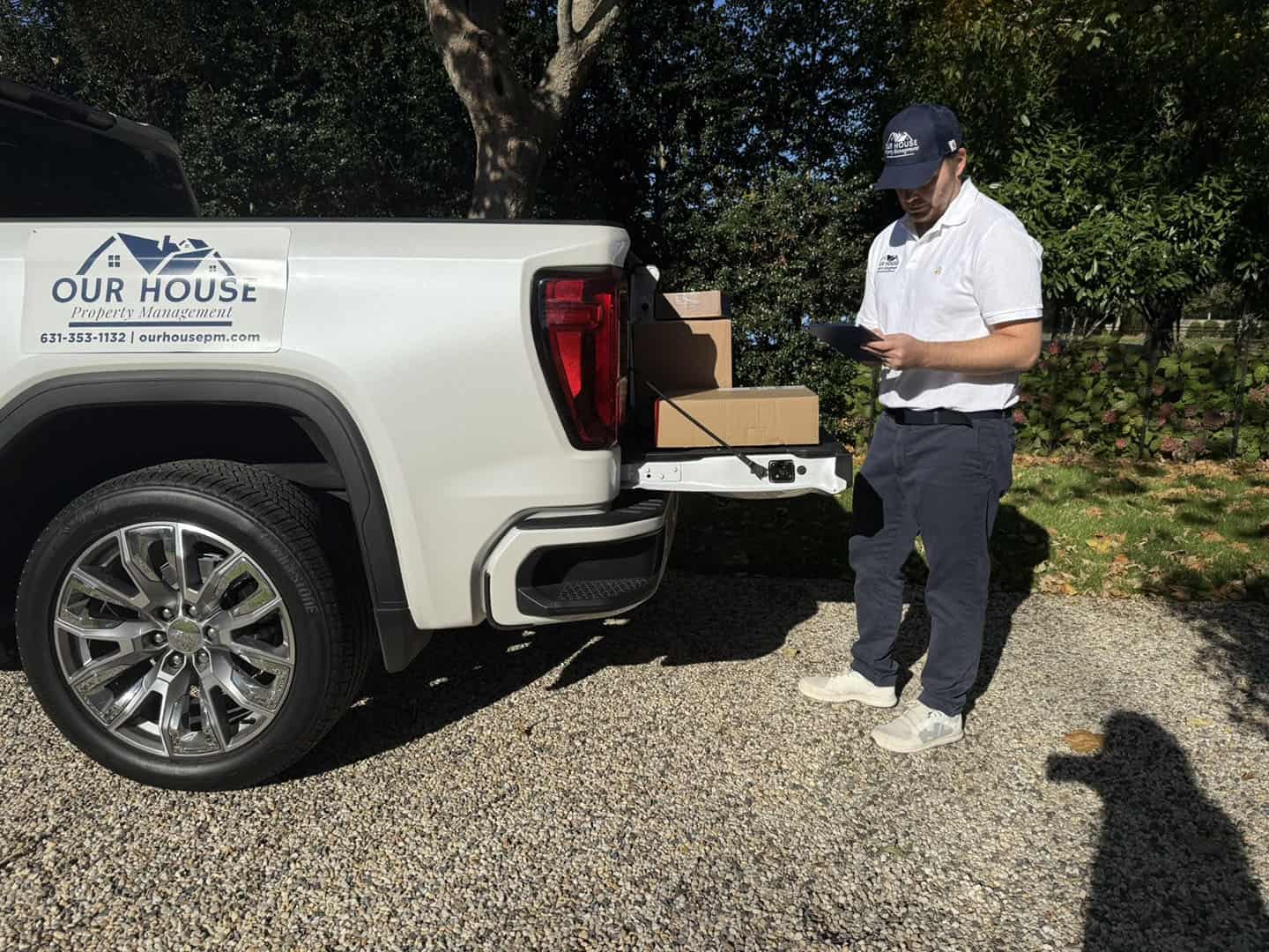 A person wearing a cap and white polo shirt stands next to an open pickup truck tailgate with boxes. The truck is branded "Our House Property Management." They are holding a tablet, standing on a gravel driveway with trees in the background.