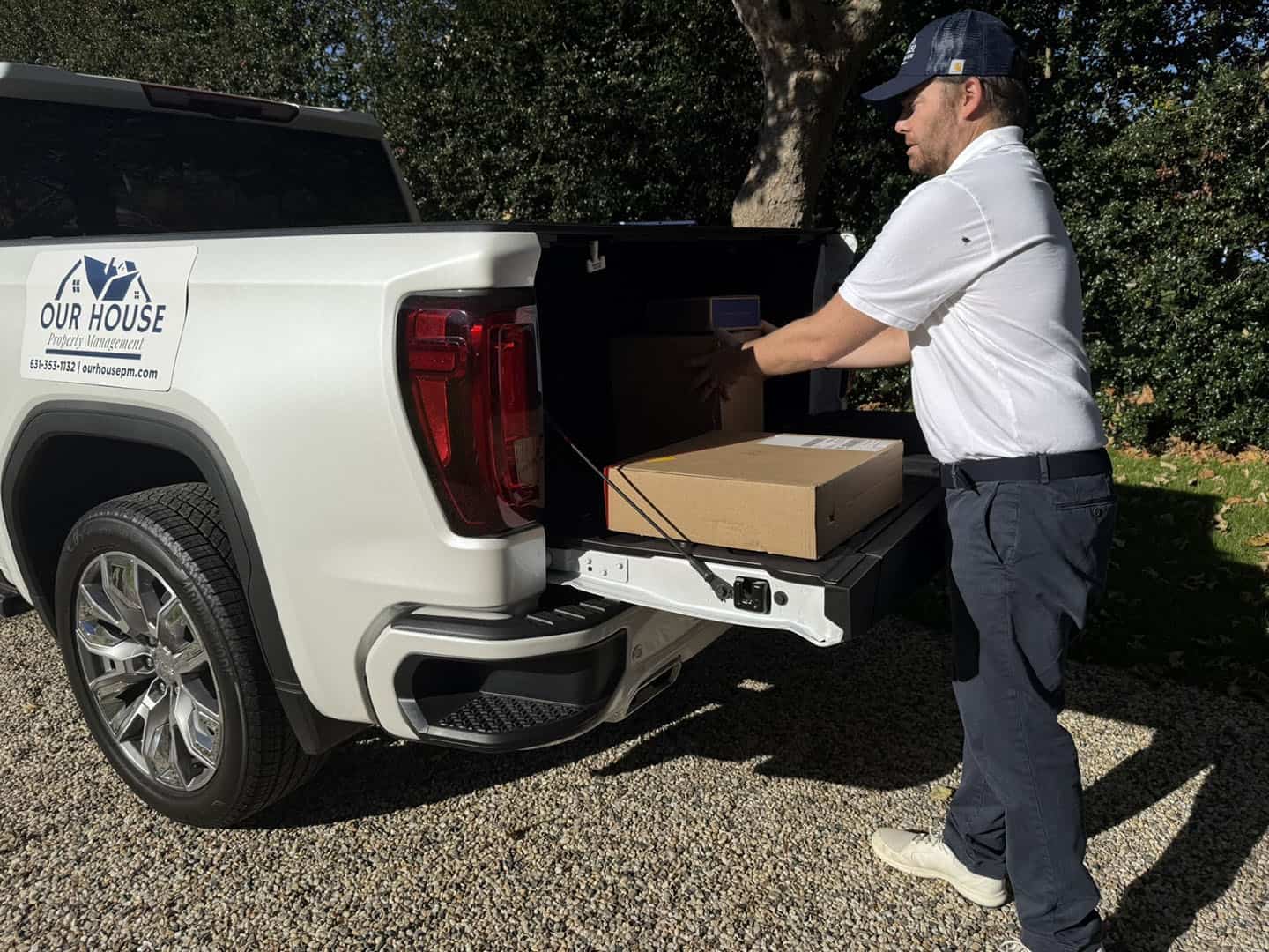 A man in a baseball cap unloads a large cardboard box from the open trunk of a white truck parked on a gravel surface. The truck has a logo on the side, and trees are visible in the background.