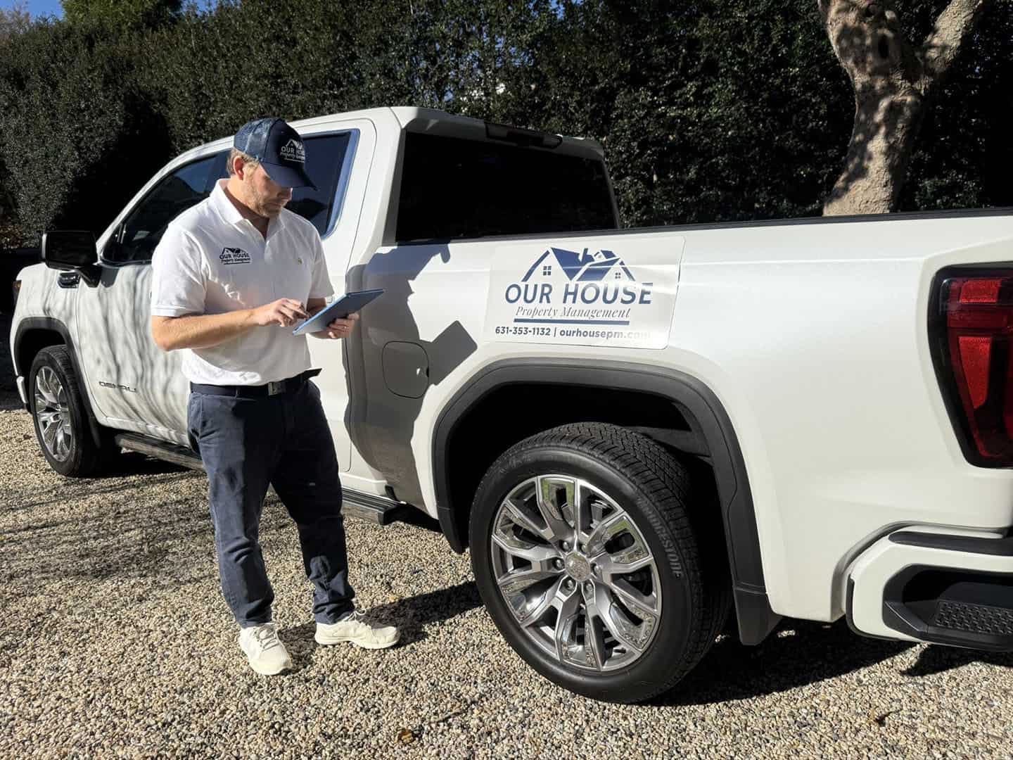 A person in a white polo and cap is using a tablet next to a white pickup truck. The truck has a sign on the side that reads "Our House Property Management" with contact details. The background features trees and a gravel surface.