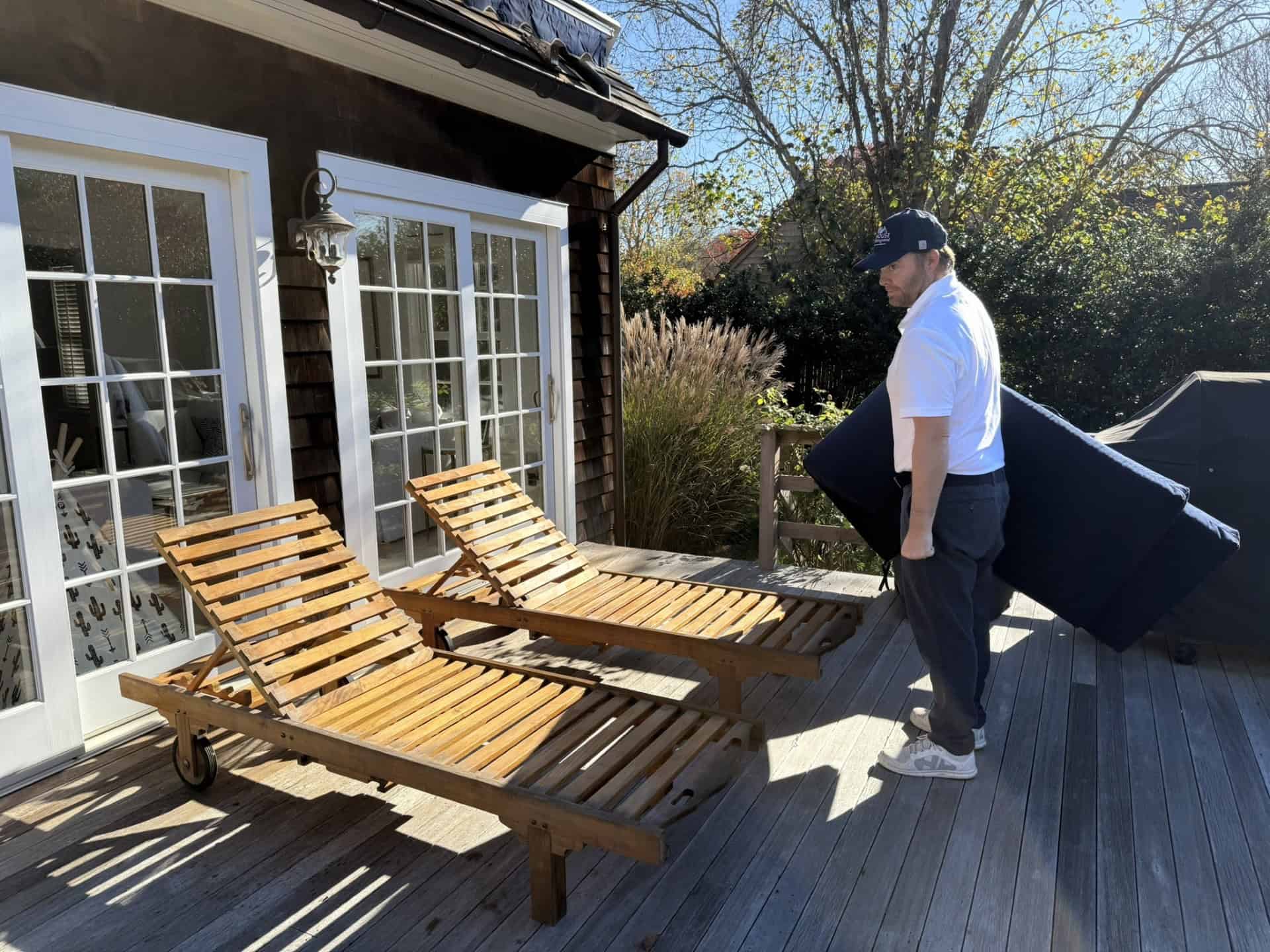 A man wearing a white shirt, cap, and pants carries a dark cushion on a wooden deck. Two wooden lounge chairs are in the foreground. Trees and a house with large windows are in the background, under a clear blue sky.