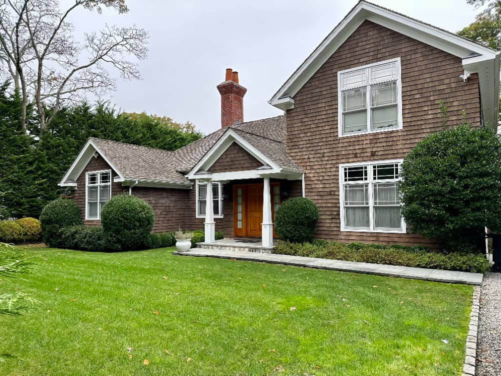 A charming two-story house with wooden shingles, large windows, and a brick chimney is ideal for rental property management. It features a front porch with white columns, surrounded by a well-maintained green lawn, bushes, and trees under a cloudy sky.