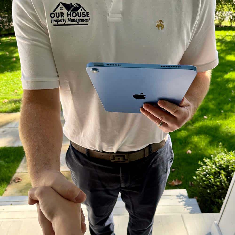 A person wearing a white polo shirt with a "Our House Property Management" logo holds a blue tablet in one hand and extends the other for a handshake. They are standing outdoors on a sunny day with a grassy background.