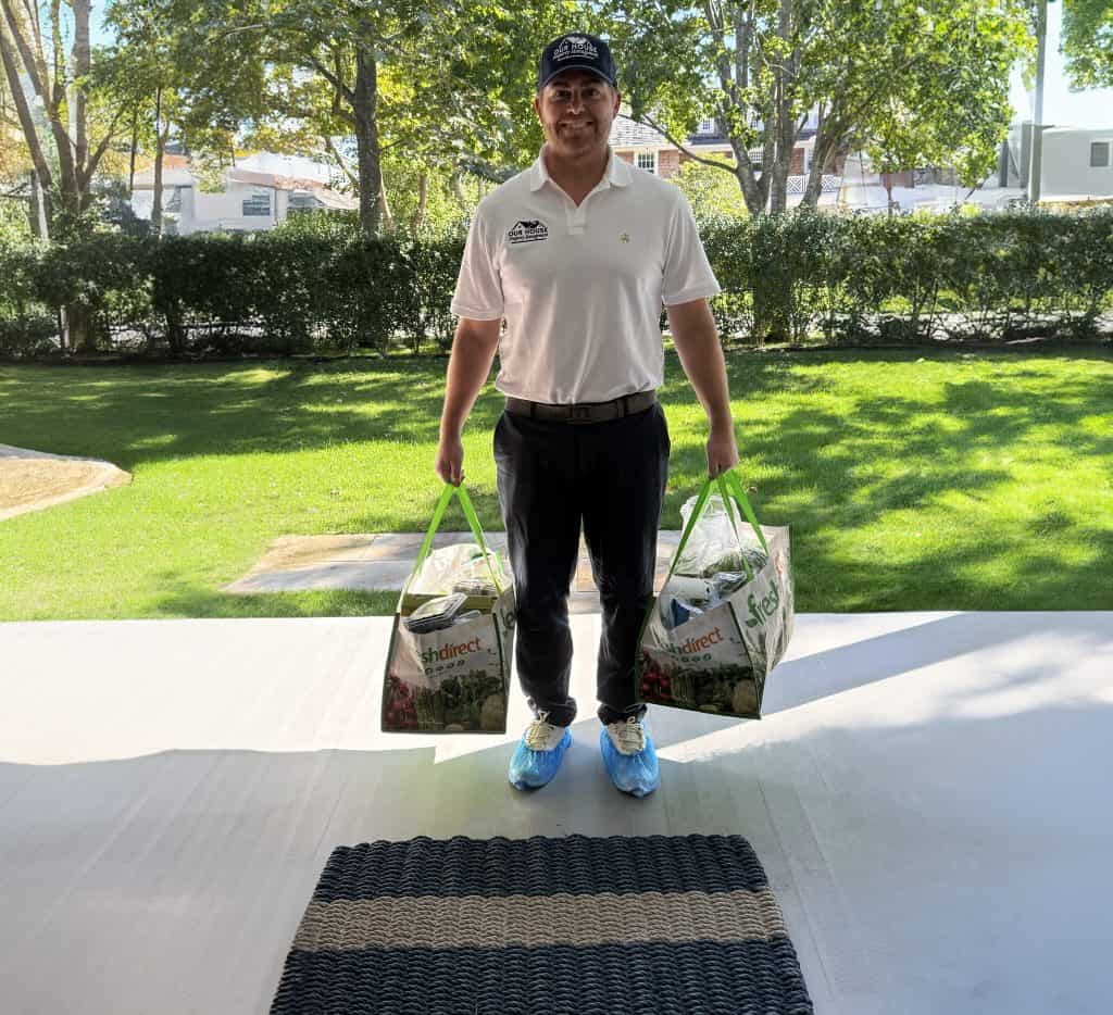 A man in a white shirt and cap stands on the porch of a rental property, holding two grocery bags. Wearing shoe covers to maintain cleanliness, he is framed by a sunny garden with green trees and grass.