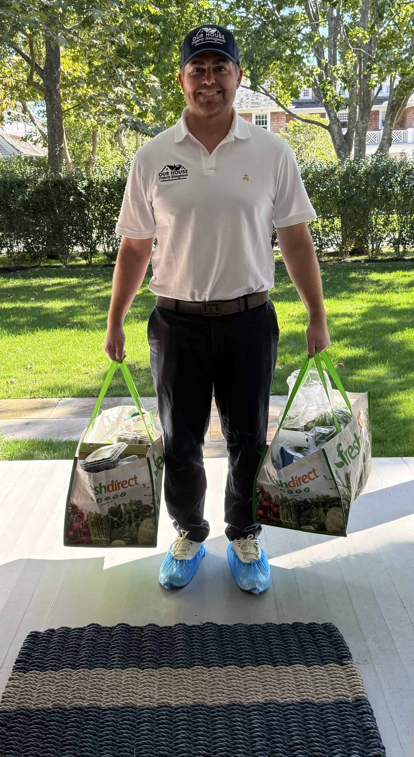 A person stands on the porch, wearing a white polo shirt, dark pants, a cap, and blue shoe covers. With an air of professionalism befitting property management, they hold two large shopping bags with green handles. Lush trees and grass create a serene backdrop.