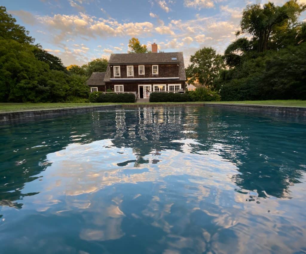 A charming countryside house with a wooden exterior, nestled amidst lush greenery. The serene swimming pool in the foreground mirrors the picturesque sky, offering a tranquil escape that embodies all you need to know about us.