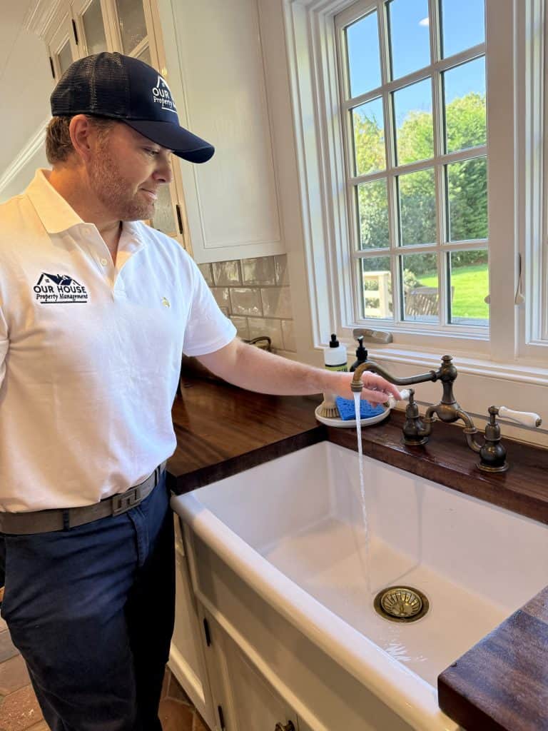 A man in a white polo shirt and dark cap stands in a kitchen managed with precision, turning on a faucet over a white sink. Sunlight streams through the window, illuminating the well-kept space. A bottle of soap sits neatly on the counter, reflecting excellent property management.