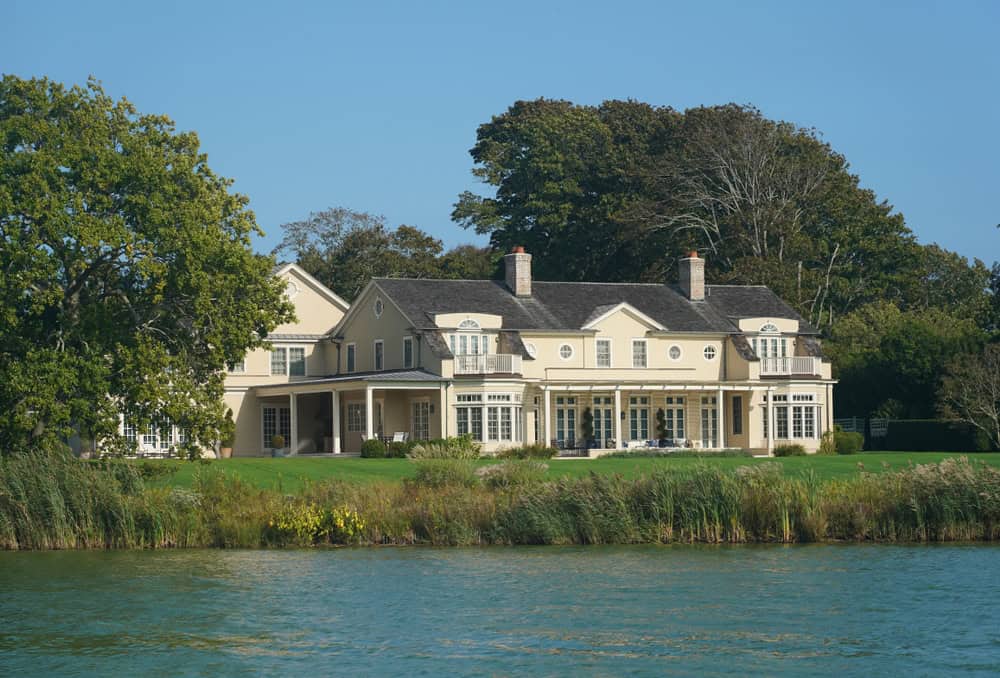 A large, elegant house with multiple chimneys and expansive windows sits near a calm body of water. Managed by a top-tier rental property management team, the property is surrounded by lush green trees and grass under a clear blue sky, ensuring both beauty and peace for its residents.