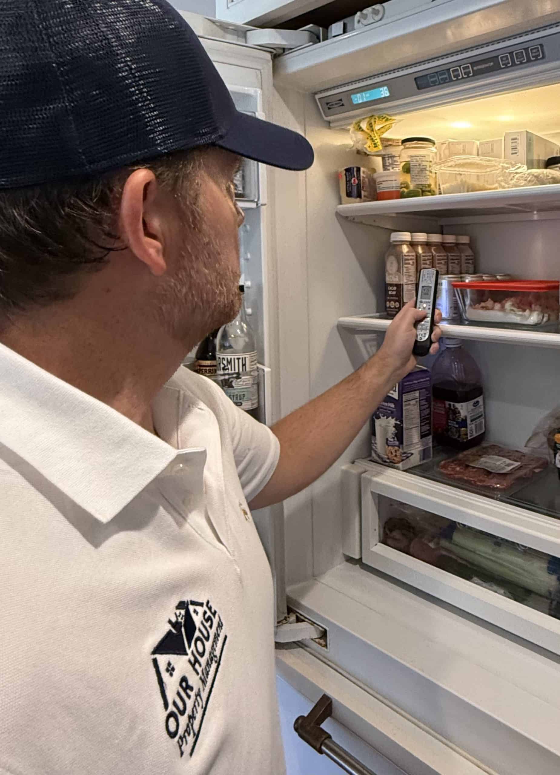 A man in a white polo and black cap, possibly tasked with rental property management duties, looks inside an open refrigerator. Holding a small item in his right hand, he examines it amidst the fridge's assortment of food and drinks.