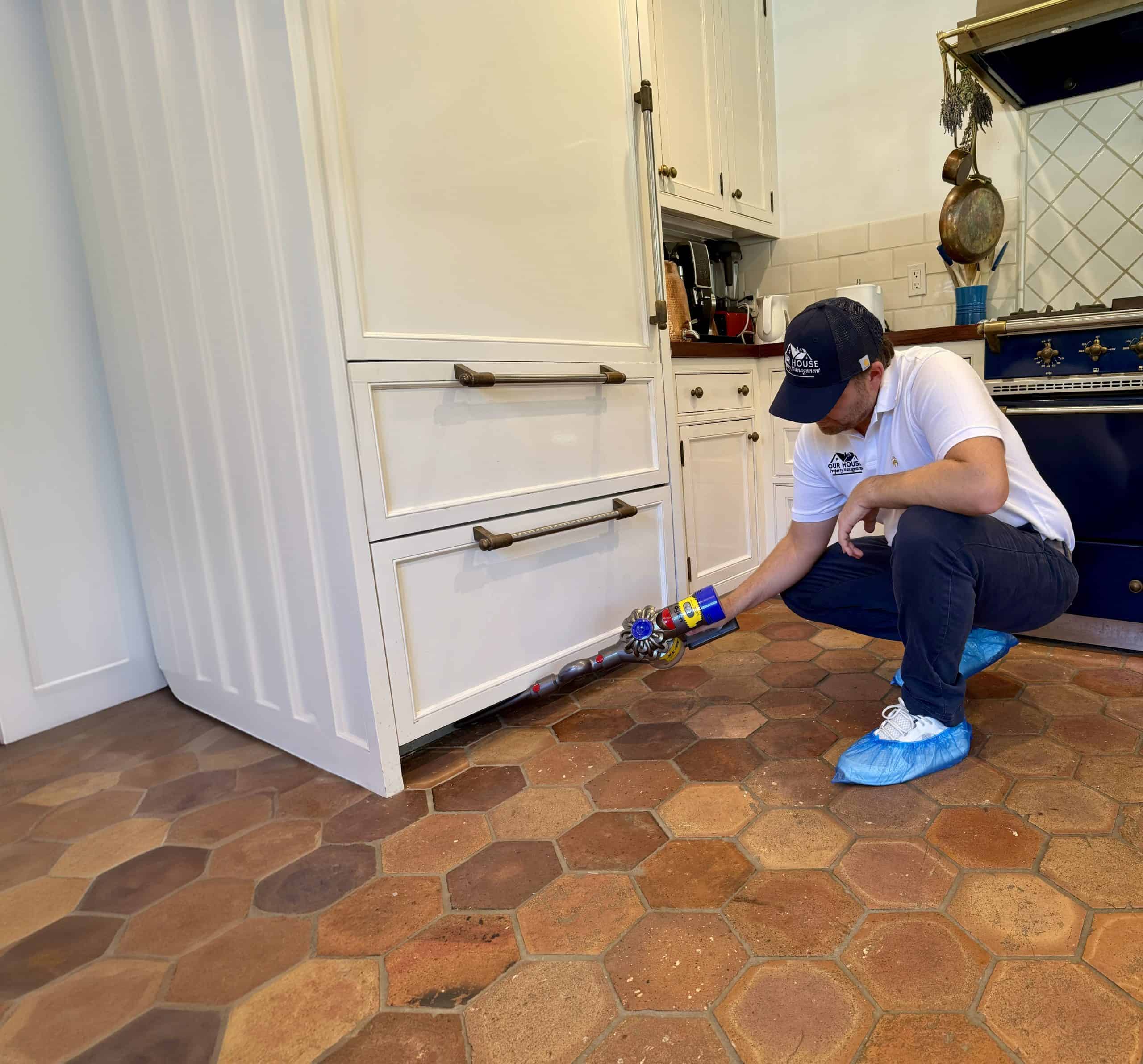 In the heart of a bustling kitchen, a person in a white shirt, cap, and shoe covers diligently tends to the base of a cabinet with a spray can. This meticulous attention to detail is crucial in rental property management, where maintaining pristine conditions is key. A blue stove and kitchen items light up the scene.