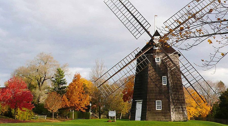 A rustic wooden windmill stands amidst colorful autumn trees with red, orange, and yellow leaves. The sky is overcast, casting a calm, serene atmosphere over the scene.