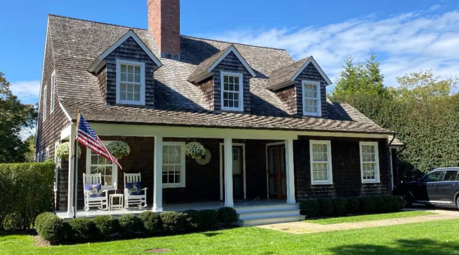 A traditional two-story house with dark wood siding and a brick chimney, expertly maintained by a dedicated property management team. It features a porch with white rocking chairs and potted plants. An American flag graces the porch, all surrounded by a well-manicured lawn under a clear blue sky.