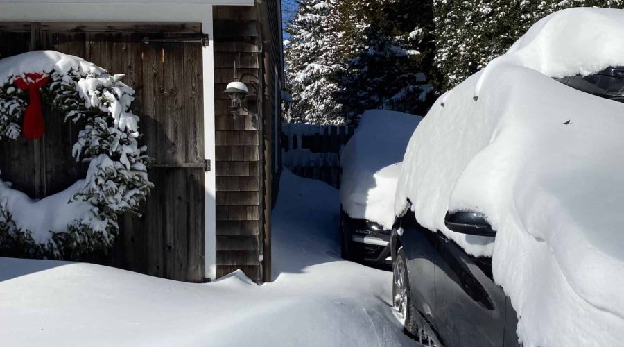 A snowy scene shows a house with a wreath, thickly covered in snow, alongside two snow-covered cars. The driveway and surroundings are blanketed in snow, with clear blue skies overhead and trees in the background.