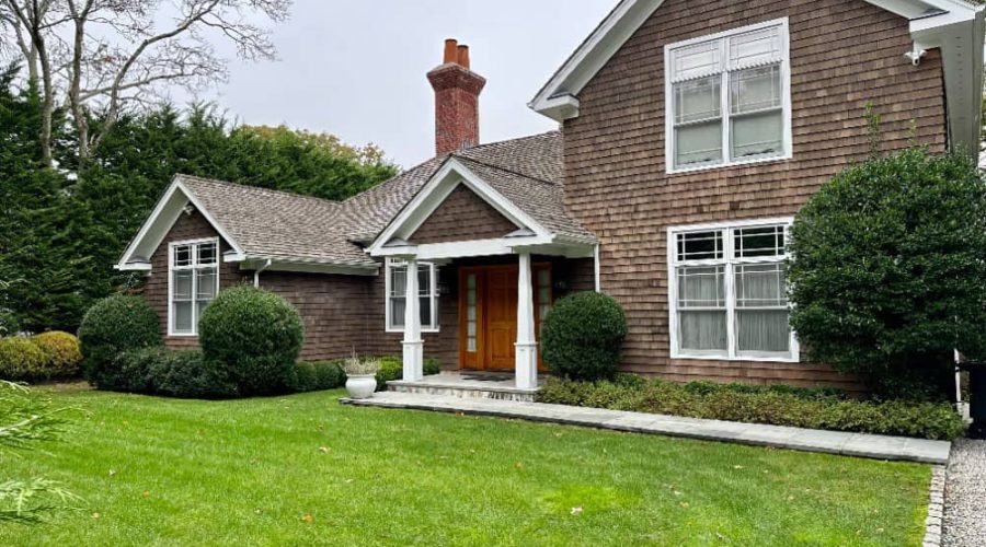 A charming two-story house with wooden shingles, large windows, and a brick chimney is ideal for rental property management. It features a front porch with white columns, surrounded by a well-maintained green lawn, bushes, and trees under a cloudy sky.