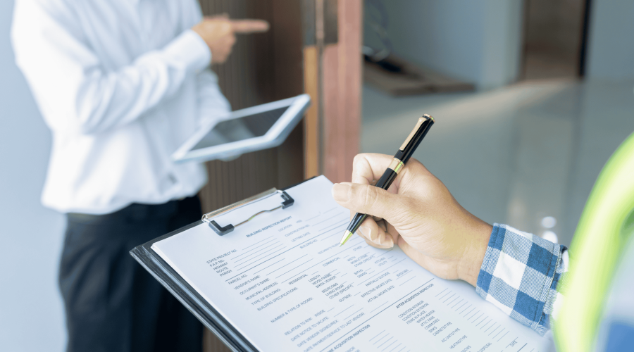 A person is holding a clipboard and pen, filling out a form in the foreground. Another person in a white shirt is holding a tablet and pointing towards a direction in the background, standing at an open door. The scene suggests an inspection or survey related to Luxury Home Management East Hampton.