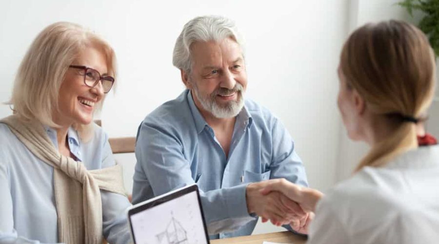 An elderly couple sits at a table with a young professional. The man shakes the professional’s hand while the woman smiles. A tablet on the table displays a building blueprint for Rental Property Management East Hampton. The scene takes place in a bright office setting.