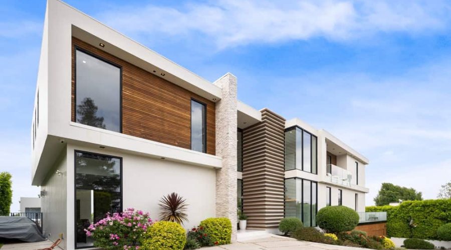 A modern two-story house with large windows and a minimalist design managed by Luxury Home Management East Hampton. The exterior showcases a mix of white and wooden panels, with a stone accent wall. The landscaped front yard features bushes and vibrant flowers under a bright blue sky.